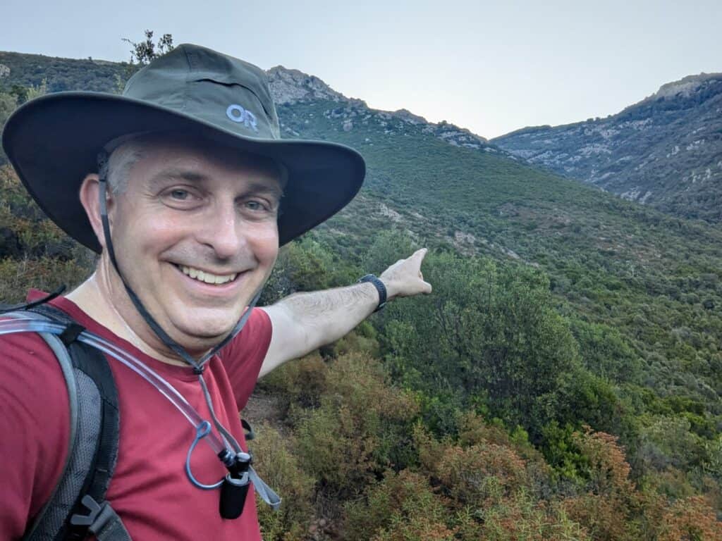 Photo of Steve pointing up the route up the mountain. The mountain is covered in green bushes. Steve is wearing a red T shirt and a green wide brimmed hat.