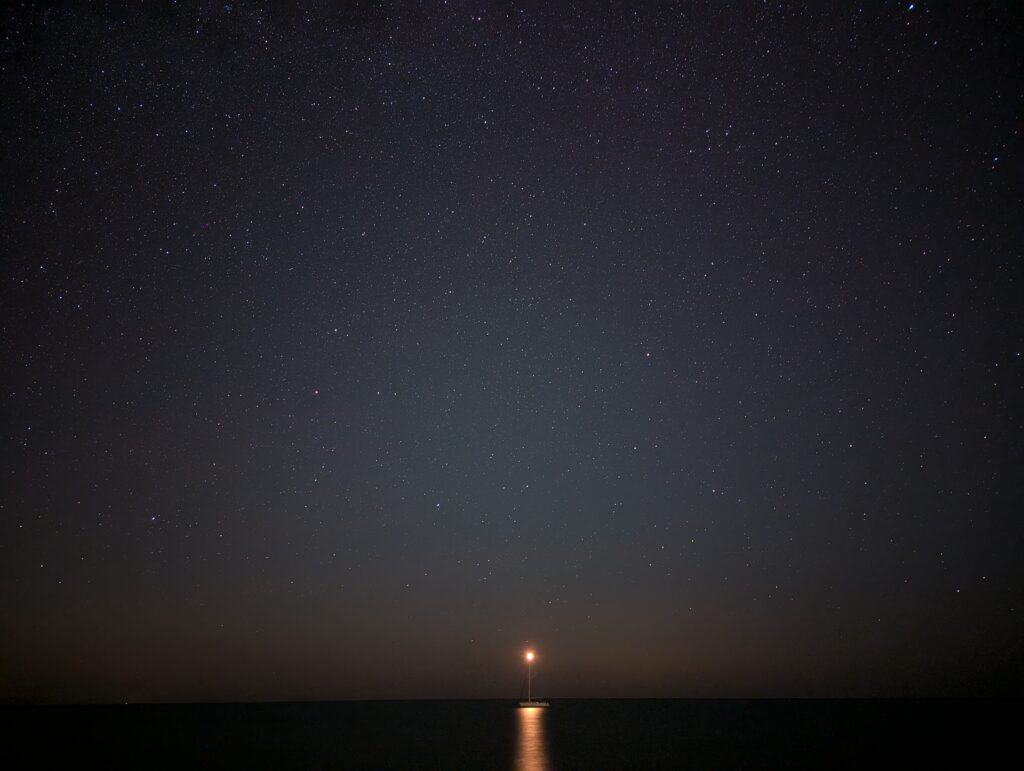 A long exposure view of the stars over the sea. A sailing boat is anchored a small way off shore with it's white anchored navigation light on top of the mast.