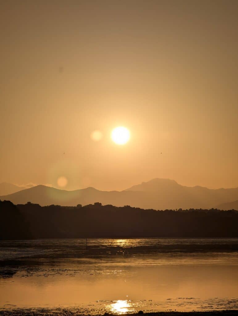 A photo of the sunset in Corsica. The sea is behind and in the photo a saltwater lake and hills that fade into mountains with the sun a little above the mountains. The colours are yellow/brown.